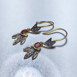 May include: A pair of gold-toned earrings with a floral design. Each earring features a central oval-shaped red gemstone, surrounded by small, clear stones. The earrings have a hook closure and a decorative element with a dark gray and silver finish.