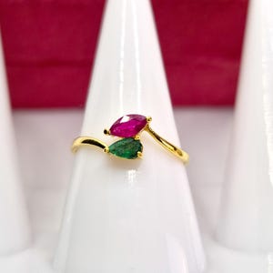 May include: A gold ring with a ruby and emerald gemstone. The ring has a pear-shaped ruby and emerald set in a delicate gold band. The gemstones are arranged to create a unique look. The ring is displayed on a white stand. The ring is a beautiful piece of jewellery.