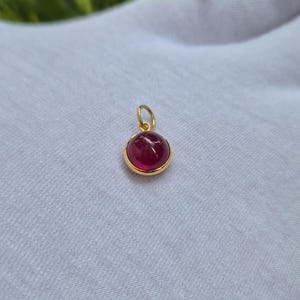 Pode incluir: Um pequeno pendente redondo com uma pedra preciosa cabochão de cor vermelho-escura e superfície lisa, engastada num bisel de metal dourado polido. O pendente inclui um pequeno anel circular dourado no topo para fixação a uma corrente.