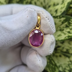 May include: An oval-shaped, faceted pink gemstone pendant set in a polished yellow gold bezel frame. The pendant features a small, integrated gold bail at the top for attachment to a chain, showcasing a vibrant, translucent pink hue and clear facets.