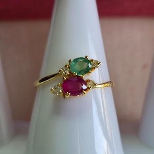 Puede incluir: Un anillo de oro con dos piedras preciosas ovaladas, una verde esmeralda y otra roja rubí. El anillo tiene pequeños detalles de diamantes a cada lado de cada piedra preciosa. El anillo se exhibe sobre una superficie blanca.