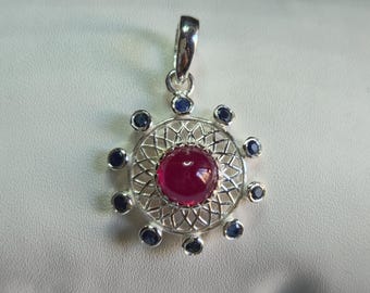May include: A silver pendant featuring a central round cabochon ruby surrounded by a delicate filigree metal lattice. The outer edge is accented with ten small, round, dark blue sapphire stones, and the piece includes a silver bail at the top.