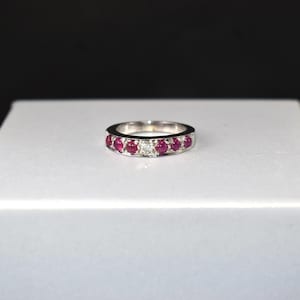 May include: A silver ring with a band of alternating red gemstones and small white gemstones. The ring is set against a white surface with a black background.