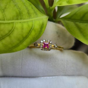 Puede incluir: Un anillo de oro con una gema roja cuadrada y vibrante en el centro, rodeada de piedras más pequeñas y transparentes. El anillo tiene piedras pequeñas y transparentes adicionales a cada lado del diseño central. El anillo está sobre un fondo de hojas verdes.