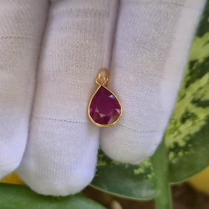 Pode incluir: Um pingente em forma de gota com uma pedra preciosa facetada em rosa escuro, engastada numa fina moldura de metal dourado. O pingente inclui um pequeno anel circular dourado na parte superior para fixação, com uma superfície polida.
