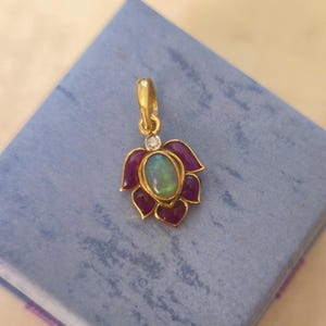 Puede incluir: Colgante de oro con diseño floral. Presenta una piedra central de ópalo ovalada, rodeada de pétalos morados oscuros. Un pequeño diamante está engastado encima del ópalo. El colgante está sobre una superficie azul.