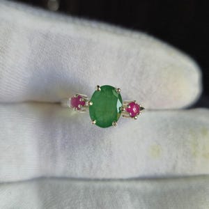 Peut inclure: Une bague en argent ornée d'une émeraude verte ovale au centre, flanquée de deux petits rubis rouges ronds. Les pierres précieuses sont serties dans une monture classique à griffes.
