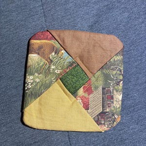 Puede incluir: Posavasos cuadrado acolchado con diseño de patchwork. El posavasos presenta una combinación de telas en tonos marrón, verde y amarillo, con un cuadrado verde central y patrones de paisaje.