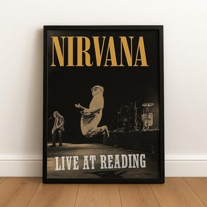 May include: Black framed poster featuring the band Nirvana. The poster has the band's name in gold lettering at the top, with the words "LIVE AT READING" at the bottom. The image shows a live performance with band members on stage.