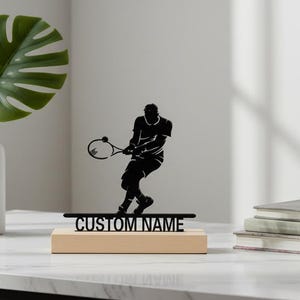 Peut inclure: Silhouette noire d'un joueur de tennis en métal sur une base en bois avec le texte "CUSTOM NAME". Une balle de tennis verte et une pile de livres sont à côté de la sculpture. Un vase blanc avec une feuille verte est à gauche.