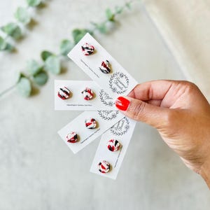 May include: Four white display cards featuring the text Ransomed Threads, each holding a pair of small stud earrings. The earrings have a marbled pattern in red, black, and white, with various shapes including hearts, circles, and squares.