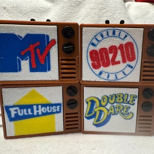 May include: Four brown plastic television-shaped keychains with white screens. The screens feature logos for MTV, Beverly Hills 90210, Full House, and Double Dare. Each keychain has two black circular knobs and a speaker grille on the right side.
