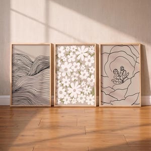 Pode incluir: Três impressões de arte emolduradas com uma estética minimalista. As impressões apresentam desenhos de linhas de montanhas, flores brancas sobre um fundo verde e um contorno de flor grande. As molduras são de madeira clara e a paleta de cores é neutra.