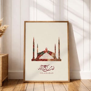 Pode incluir: Impressão artística emoldurada com uma silhueta estilizada de uma mesquita em tons de vermelho, verde e creme. A palavra "Islamabad" está impressa abaixo em vermelho escuro. Fundo creme, moldura de madeira.