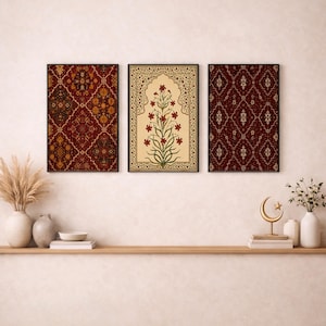 May include: A set of three rectangular wall art prints in black frames. The left and right prints feature a dark red diamond pattern with intricate gold and cream details. The center print displays a red floral plant inside a cream archway.