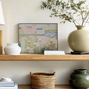 May include: A framed landscape painting depicts a pink building and white structure near a body of water, with a field of white and pink flowers in the foreground. A white vase, a large green vase, and a woven basket are also visible.