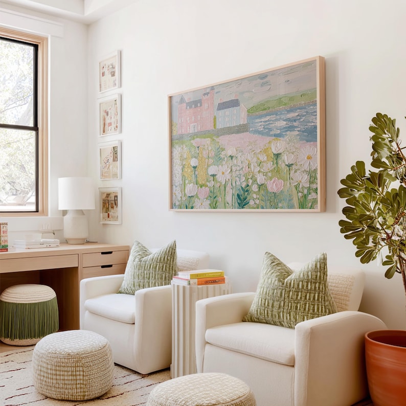 May include: A bright, airy living space with white armchairs, green patterned pillows, and a large framed painting. The painting depicts a landscape with a pink building and white flowers. A desk with a lamp and a fringed ottoman are also visible.