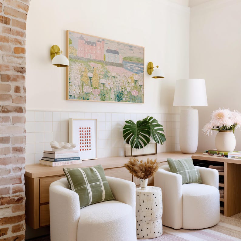 May include: A cozy living room scene featuring two white, curved armchairs with green plaid pillows. A landscape painting hangs above a wooden cabinet. Decorative elements include a white lamp, a vase of flowers, and a side table with a speckled design.