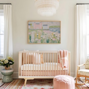 May include: A light wood crib with a pink mattress and bedding, a pink blanket, and a pink ottoman. A framed painting of a landscape with a pink building hangs above the crib. The room has white curtains and a chandelier.