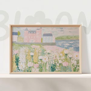 May include: A framed landscape painting featuring a coastal scene with a pink castle, blue houses, and a field of white and pink flowers. The artwork is set against a backdrop of a blue sea and sky, with a light wooden frame.