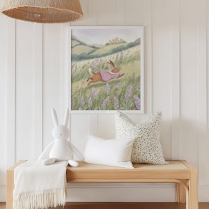 May include: A framed watercolor print of a running rabbit wearing a pink cape in a field of purple flowers. A white plush bunny, two pillows, and a fringed blanket sit on a wooden bench. A woven pendant light hangs above.
