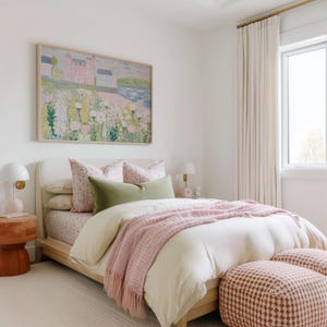 May include: A bedroom scene featuring a bed with a light wood frame, adorned with floral print pillows, a green lumbar pillow, and a pink fringed throw. A framed landscape painting hangs above the bed. Two patterned ottomans sit at the foot of the bed.