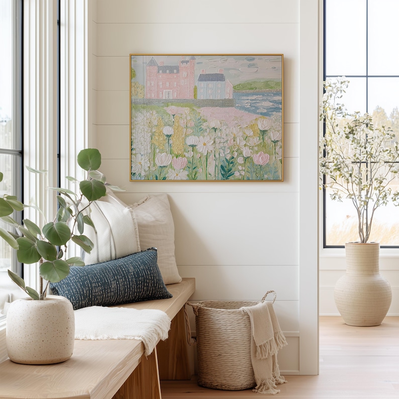 May include: A cozy interior scene featuring a framed landscape painting with a pink and blue castle. A wooden bench with pillows and a woven basket with a blanket are in the foreground. A large vase with a plant sits in the background.
