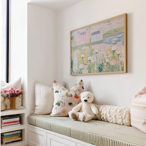 May include: A cozy window seat with a green and white striped cushion, adorned with decorative pillows and a teddy bear. A framed painting of a pink building and flowers hangs above. A shelf displays books and a vase of pink flowers.