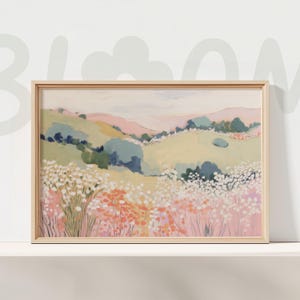 May include: A framed landscape art print featuring a pastel color palette. The artwork depicts rolling hills in shades of pink, yellow, and green, with clusters of trees and wildflowers in the foreground. The frame is a light wood color, and the overall style is soft and inviting.
