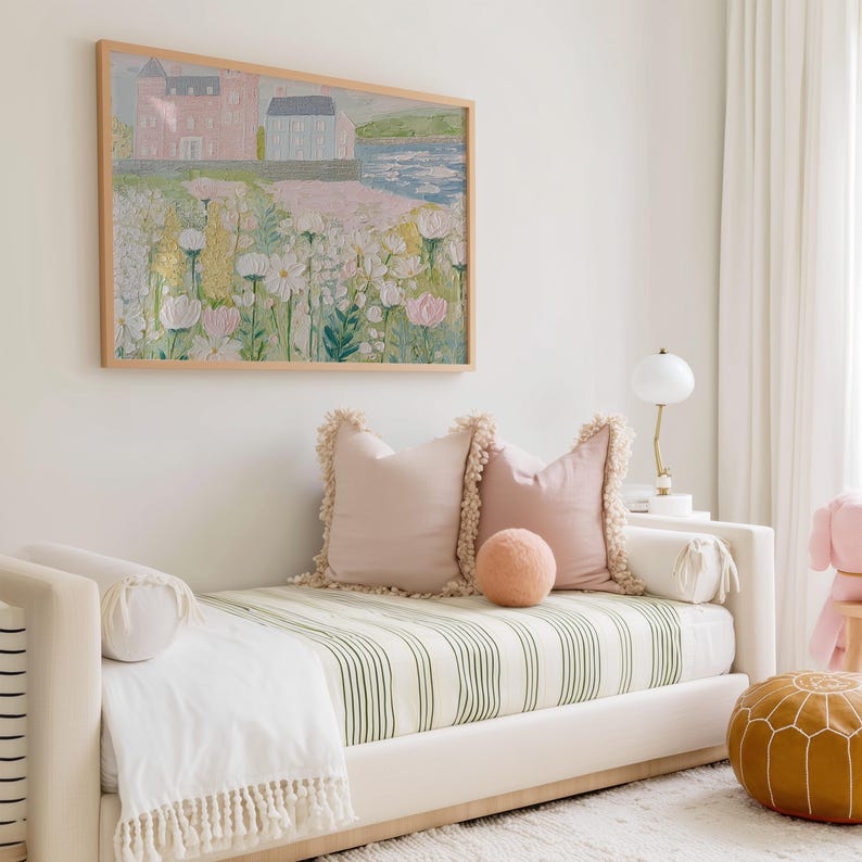 May include: A light-colored daybed with a green and white striped mattress is adorned with pink pillows and a round peach-colored accent pillow. A framed landscape painting hangs above the bed, and a small lamp sits on a side table.