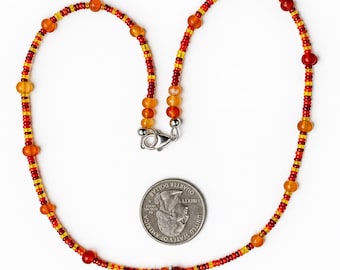 May include: A beaded necklace featuring small red, orange, and yellow seed beads interspersed with larger round orange and red beads. A silver-toned teardrop-shaped pendant with an intricate filigree pattern hangs from the center. A US quarter is shown for scale.