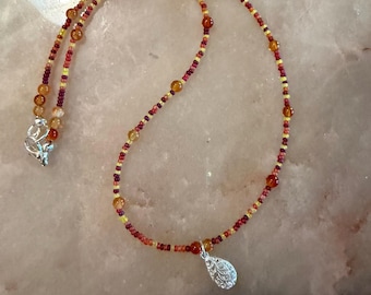 Puede incluir: Un collar de cuentas con una mezcla de pequeñas semillas de color amarillo, naranja y rojo oscuro, intercaladas con cuentas más grandes y translúcidas en tonos ámbar. Un colgante plateado en forma de lágrima con un patrón en relieve.