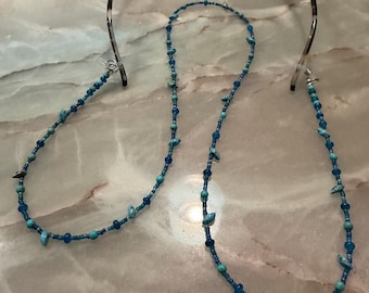 May include: A pair of tortoise shell eyeglasses attached to a long beaded chain. The chain features a variety of small blue and turquoise beads in different shapes and sizes, connected by clear silicone loops at each end of the frames.