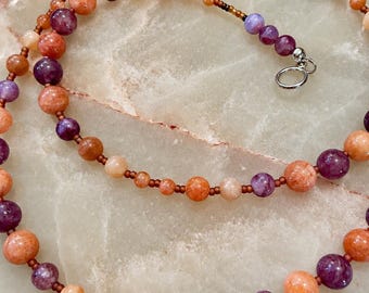 Puede incluir: Un collar de cuentas con cuentas redondas alternas en tonos naranja, melocotón y morado intenso. Las cuentas están ensartadas en un cordón fino con pequeñas cuentas espaciadoras marrones, terminado con un cierre circular plateado.