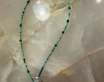 May include: A delicate beaded necklace featuring small turquoise and green gemstone beads interspersed with tiny dark blue accents. The necklace includes a silver-toned metal extender chain with circular links and a lobster claw clasp closure.