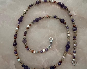 May include: A beaded necklace featuring a mix of round purple amethyst beads, small tan and brown earth-toned beads, and white speckled beads. The strand is arranged in a spiral pattern and secured with a silver-toned metal lobster claw clasp.