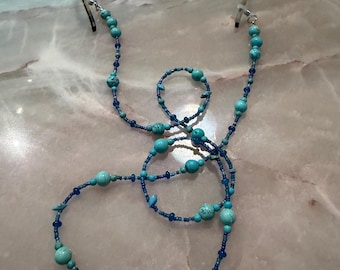 May include: A glasses chain featuring a series of turquoise-colored stone beads in varying sizes interspersed with small, translucent dark blue glass beads. The chain is attached to the temples of a pair of tortoiseshell-patterned eyeglasses.