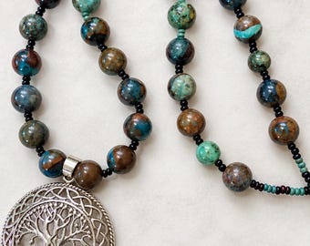 May include: A necklace featuring a large, round, silver-toned metal pendant with an intricate tree of life design enclosed in a braided border. The strand consists of variegated blue, green, and brown stone beads interspersed with small black beads.