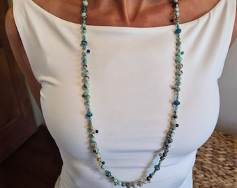 May include: A long necklace featuring a variety of round beads in shades of turquoise, teal, blue, and grey. The beads are interspersed with small metallic accents and tiny dark blue glass beads, creating a multi-textured, beaded strand design.