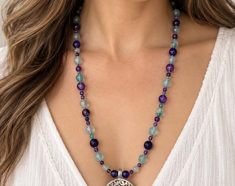 May include: A necklace featuring a circular silver-toned pendant with an intricate tree design. The chain consists of alternating round beads in shades of deep purple, translucent light blue, and dark blue, creating a colorful beaded pattern.