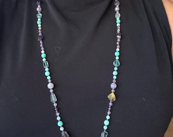 May include: A long beaded necklace featuring a mix of round and irregularly shaped beads in shades of purple, teal, light green, and yellow. The beads are strung together in a varied pattern, creating a colorful accessory that hangs down.