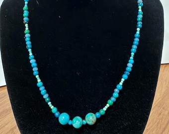 May include: A beaded necklace featuring a variety of round beads in shades of turquoise, teal, and light blue. The strand is composed of smaller beads that gradually increase in size toward the center, where three larger, prominent beads are placed.