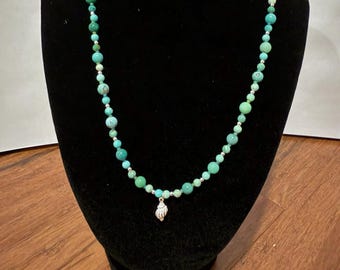 May include: A beaded necklace featuring various shades of green and turquoise round beads interspersed with small silver-toned spacers. A single silver-toned seashell charm hangs from the center of the strand, creating a focal point.