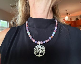 May include: A necklace featuring a circular silver-toned pendant with a tree of life cutout design, suspended from a strand of multicolored, smooth, round beads in shades of pink, purple, lavender, and light blue, worn against a black top.