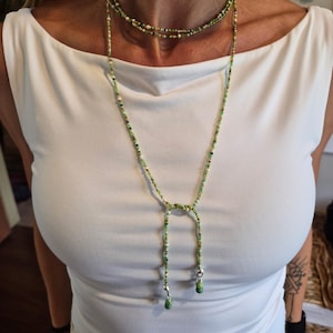 May include: A long, multi-strand beaded necklace featuring small glass beads in various shades of green, white, and gold. The necklace wraps around the neck and features a lariat-style drop with two dangling ends, each finished with a larger bead.