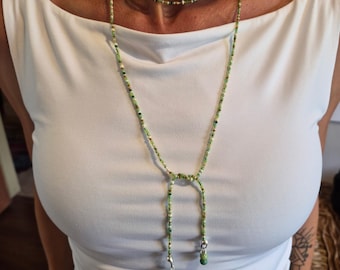 May include: A long, multi-strand beaded necklace featuring small glass beads in various shades of green, white, and gold. The necklace wraps around the neck and features a lariat-style drop with two dangling ends, each finished with a larger bead.