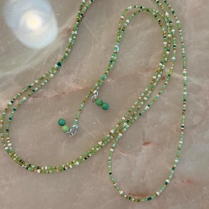 May include: A long beaded necklace featuring a mix of small green, white, and metallic gold seed beads. The strand is accented with two small, round, turquoise-coloured stone beads at the ends, which are attached with silver-toned metal clasps.