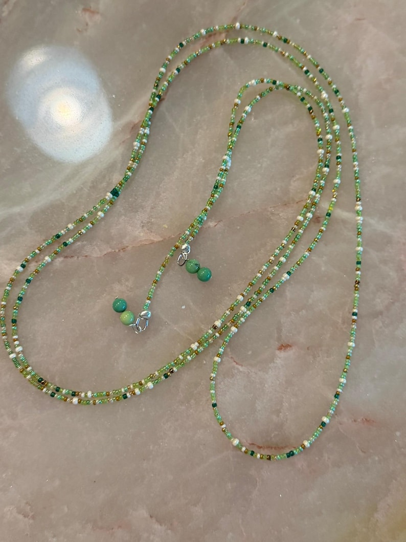 May include: A long beaded necklace featuring a mix of small green, white, and metallic gold seed beads. The strand is accented with two small, round, turquoise-coloured stone beads at the ends, which are attached with silver-toned metal clasps.