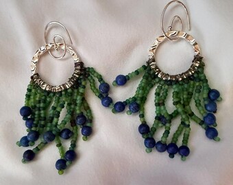 May include: A pair of dangle earrings featuring silver-toned hammered circular hoops. Attached to the bottom of each hoop are multiple strands of small green seed beads interspersed with round, dark blue beads, creating a fringe effect.