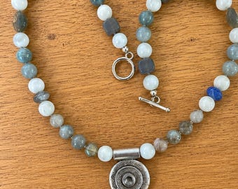 May include: A beaded necklace featuring a mix of round white, grey, and blue-grey beads, including some square-shaped grey beads. The necklace includes a silver-toned toggle clasp and a circular silver-toned pendant with an etched pattern.